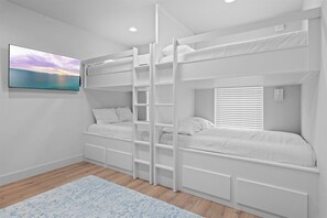 A bunk bed room with a ladder, a window with blinds, a wall-mounted TV, and a patterned rug.