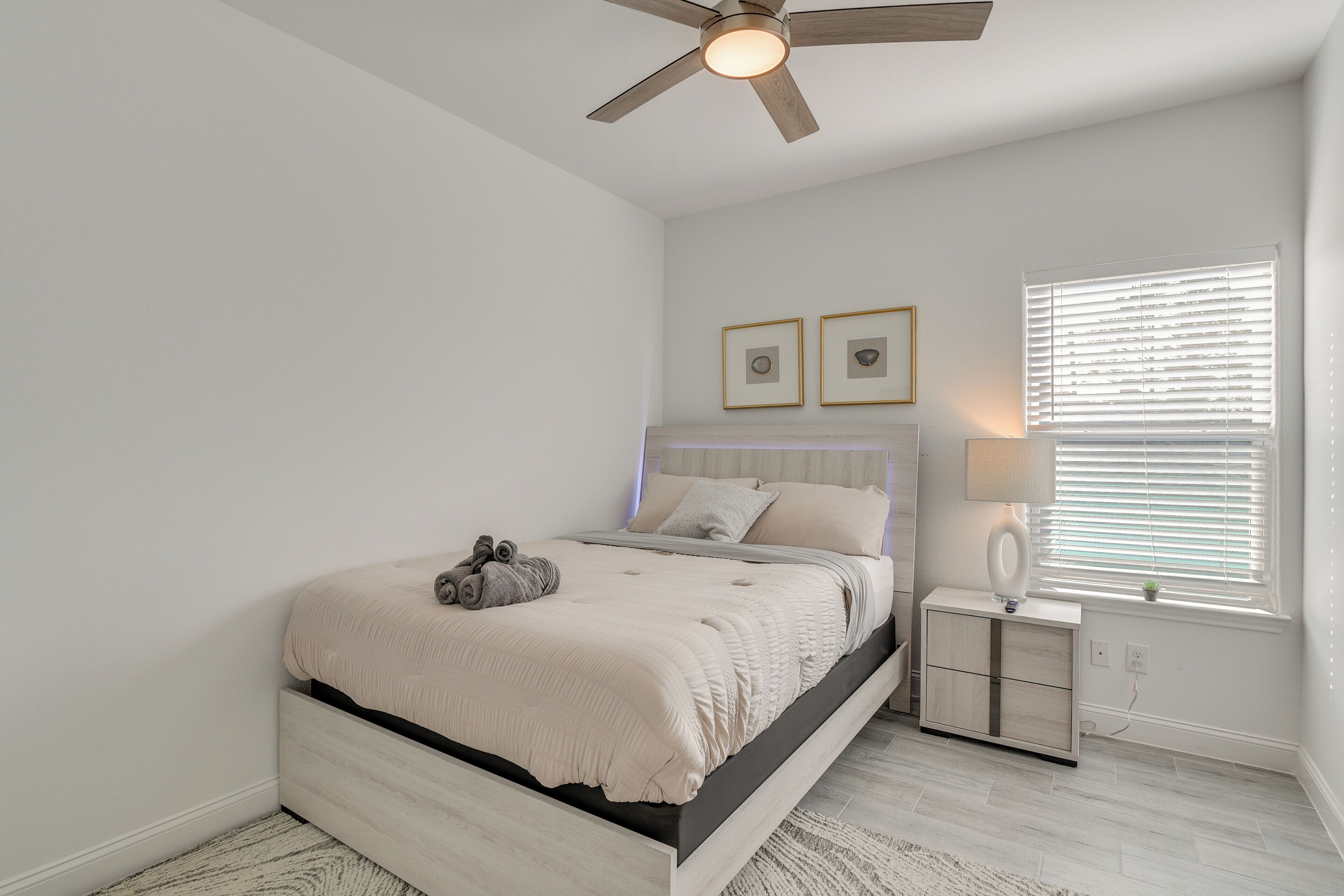 A bedroom with a bed, a nightstand, a ceiling fan, a window with blinds, and two framed pictures on the wall.