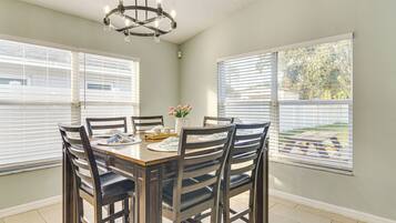 A dining area with a wooden table and chairs, a chandelier, and large windows with blinds.