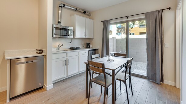 Cocina compacta con armarios blancos, microondas integrado y lavavajillas. Hay una mesa de comedor con cuatro sillas y una ventana con cortinas.