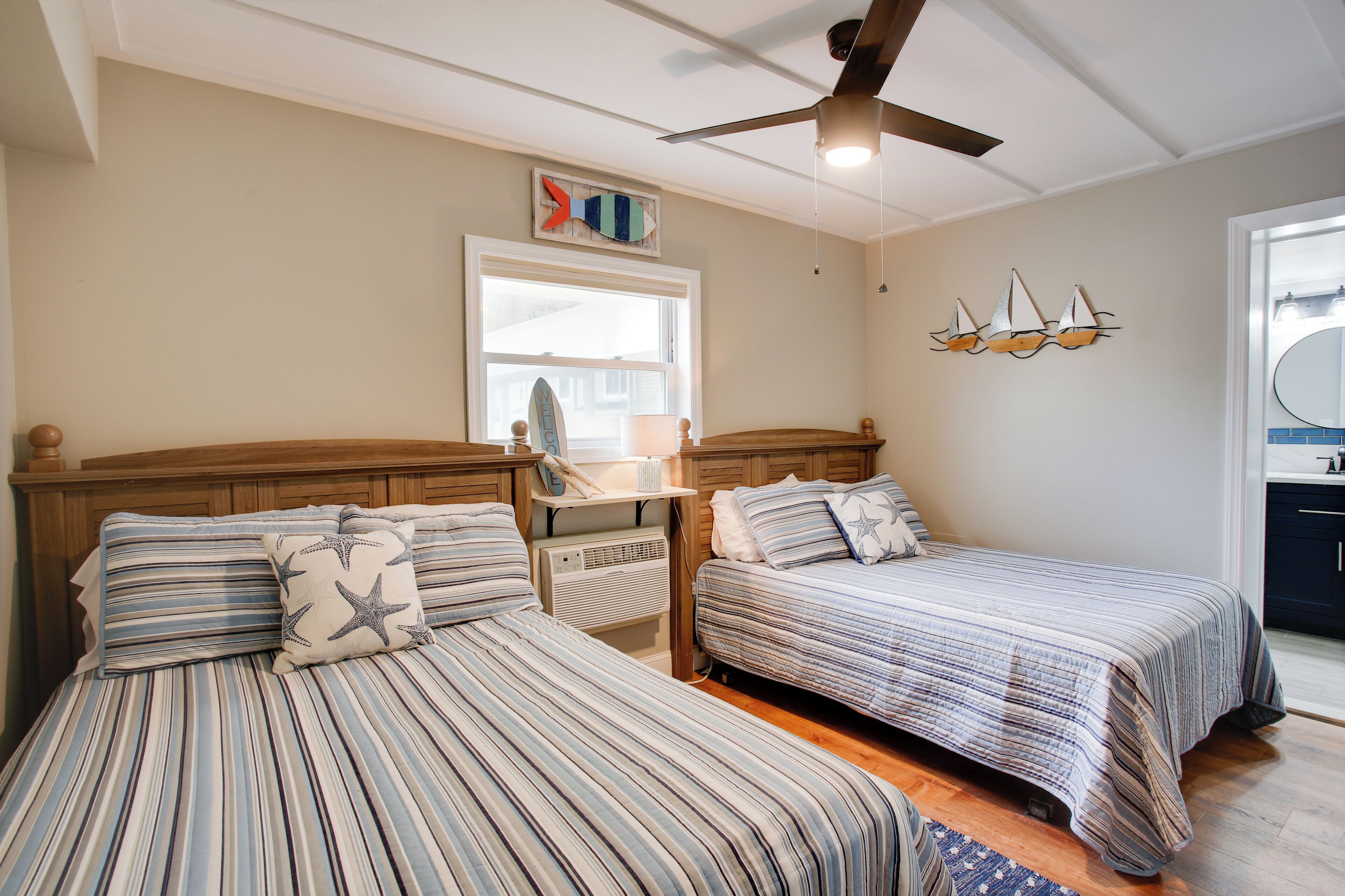 A bedroom with two beds, a ceiling fan, a window, and a wall decoration of sailboats.