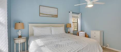 A bedroom with a bed, two nightstands, a ceiling fan, a window with curtains, and a wall-mounted picture.