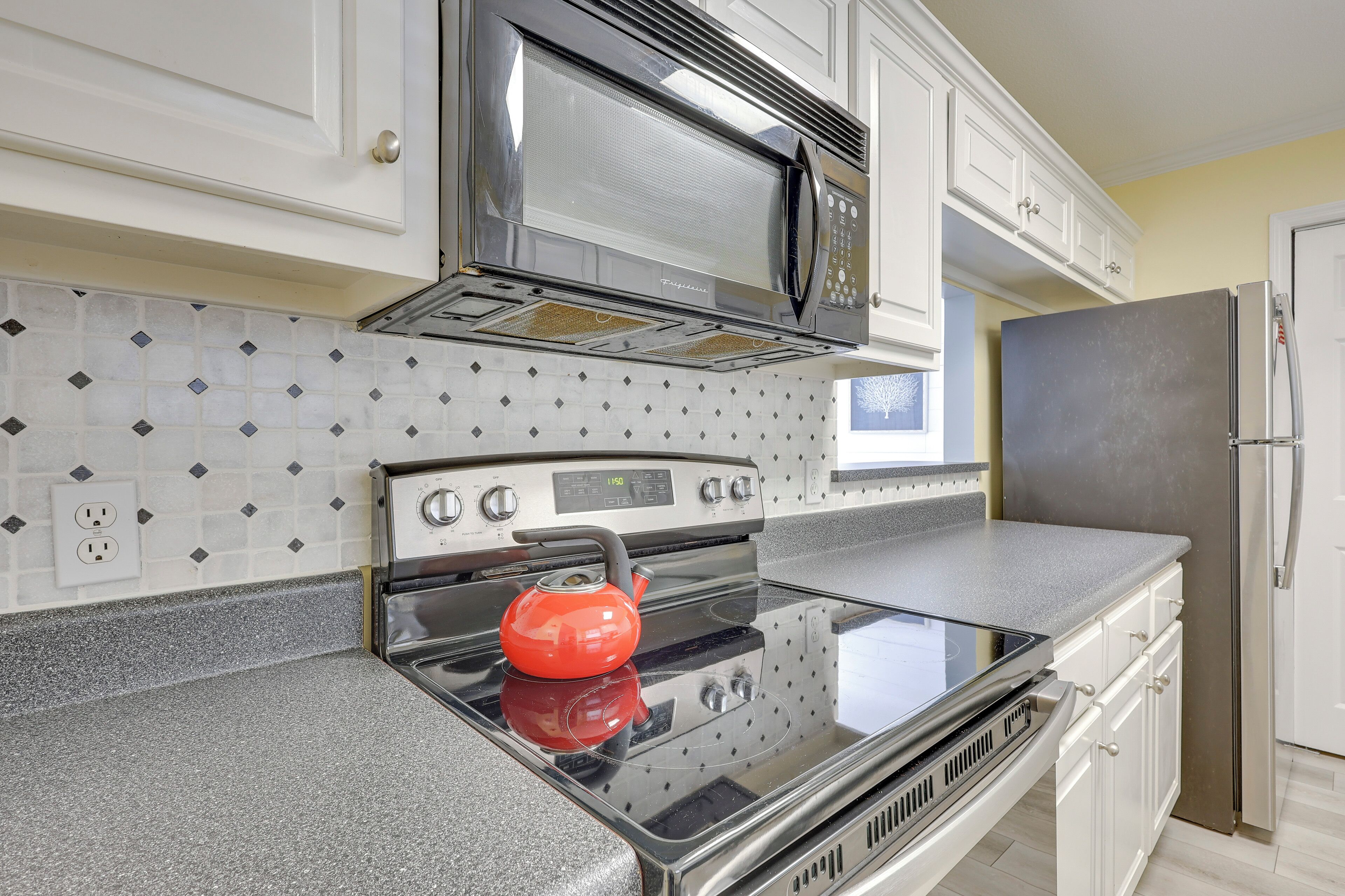 A kitchen with white cabinets, a stainless steel microwave, a stove with a kettle, and a refrigerator.