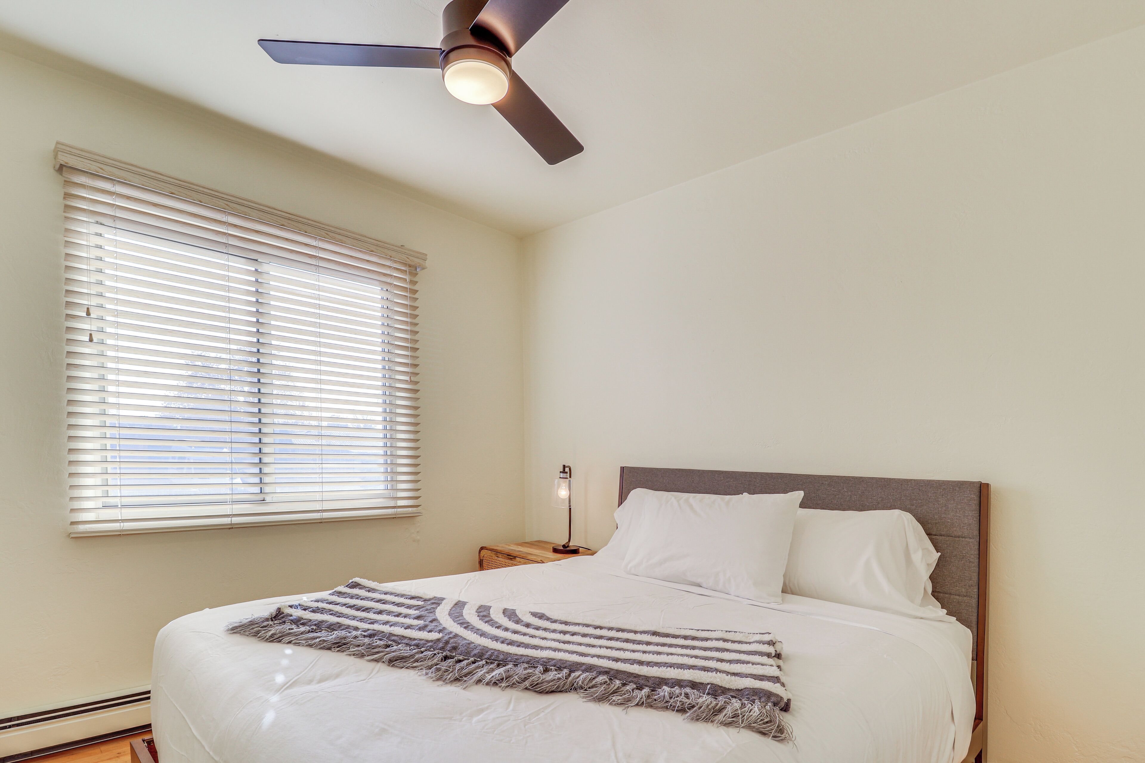 Un dormitorio con cama, ventilador de techo, mesita de noche, lámpara y ventana con persianas.