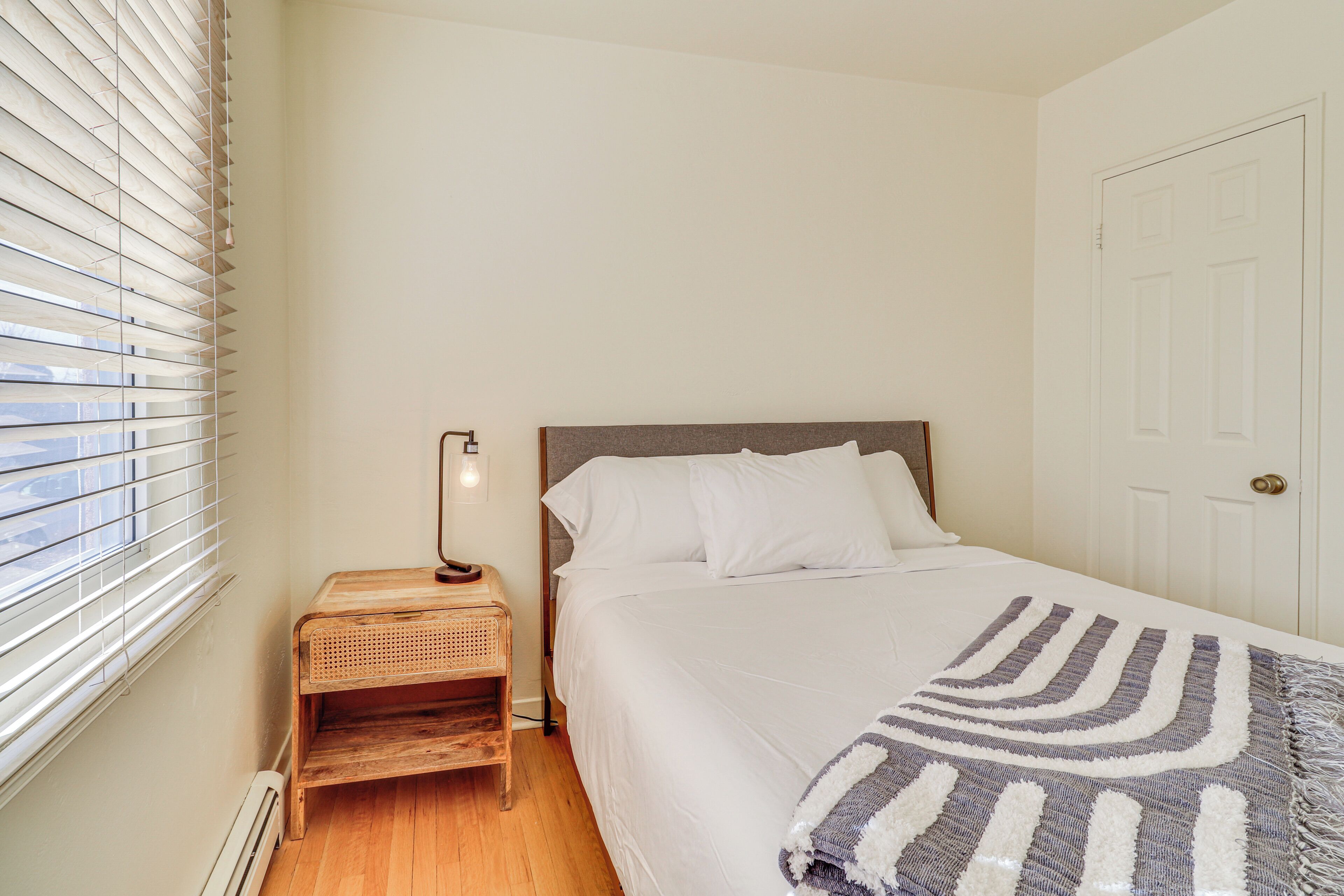 A bedroom with a bed, a bedside table with a lamp, a window with blinds, and a door.
