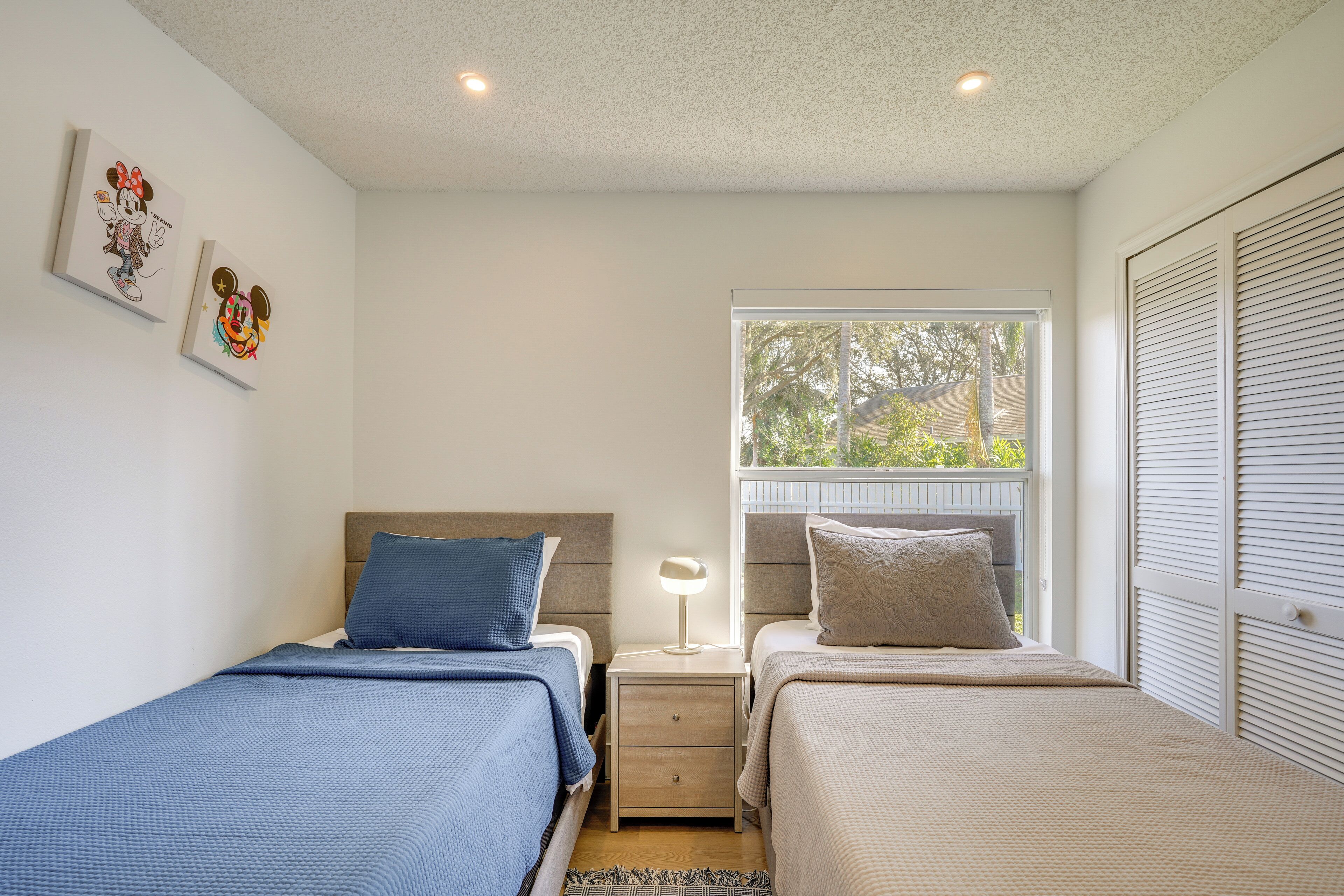 A bedroom with two beds, a nightstand with a lamp, and a window with a view of greenery.