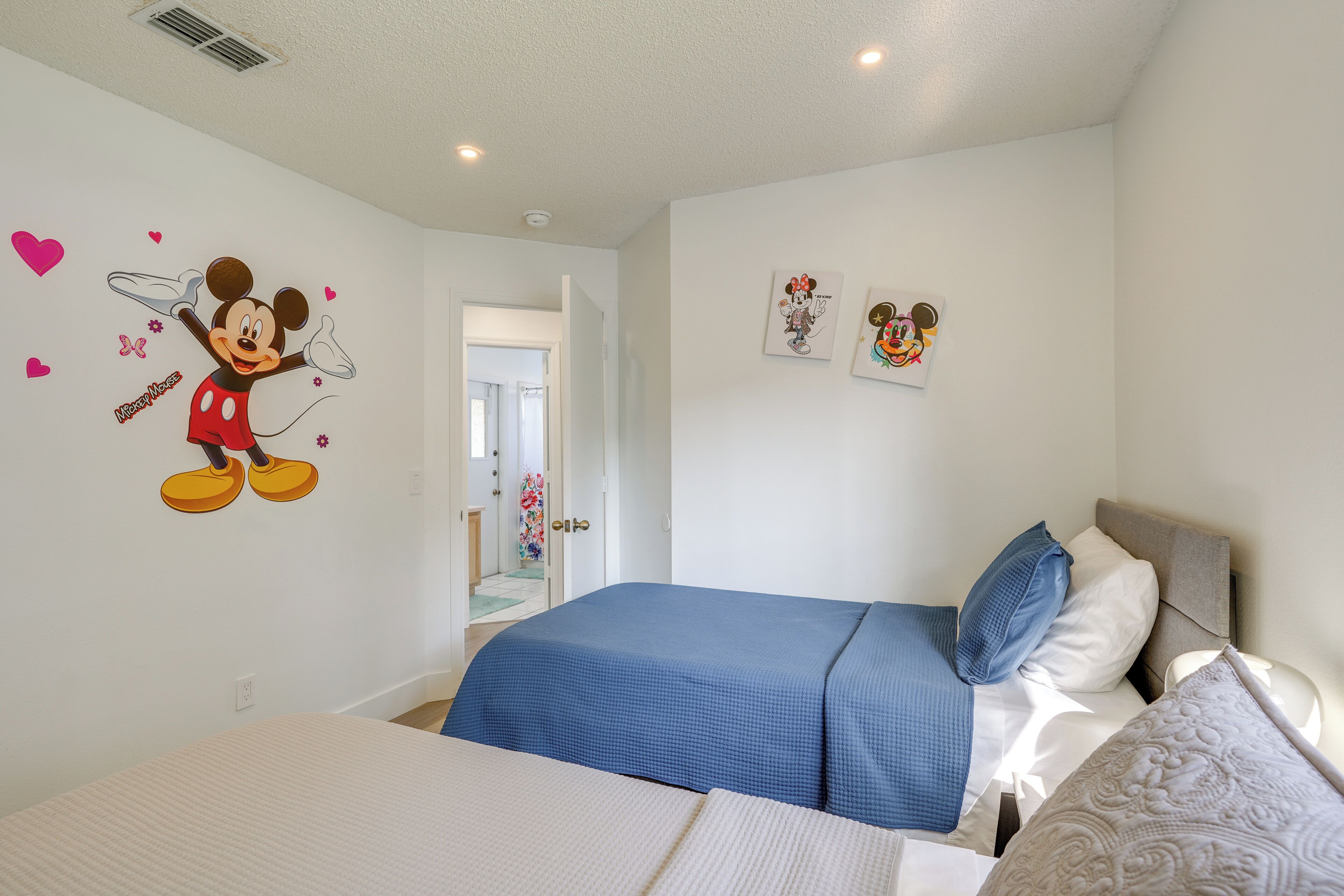 A bedroom with a bed, a blue bedspread, and a Mickey Mouse wall decal.
