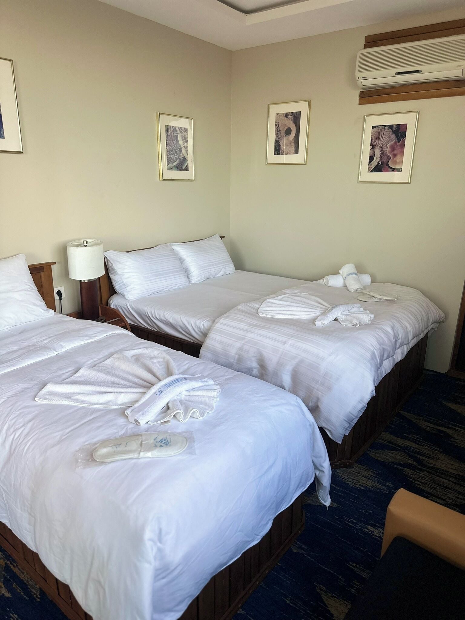 Habitación de hotel con dos camas, cada una con ropa de cama y toallas blancas. Hay cuadros enmarcados en la pared, una lámpara en la mesita de noche y un equipo de aire acondicionado sobre las camas.