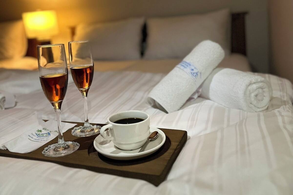 A neatly made bed with a rolled towel, two glasses of a dark beverage, and a cup of coffee on a tray.