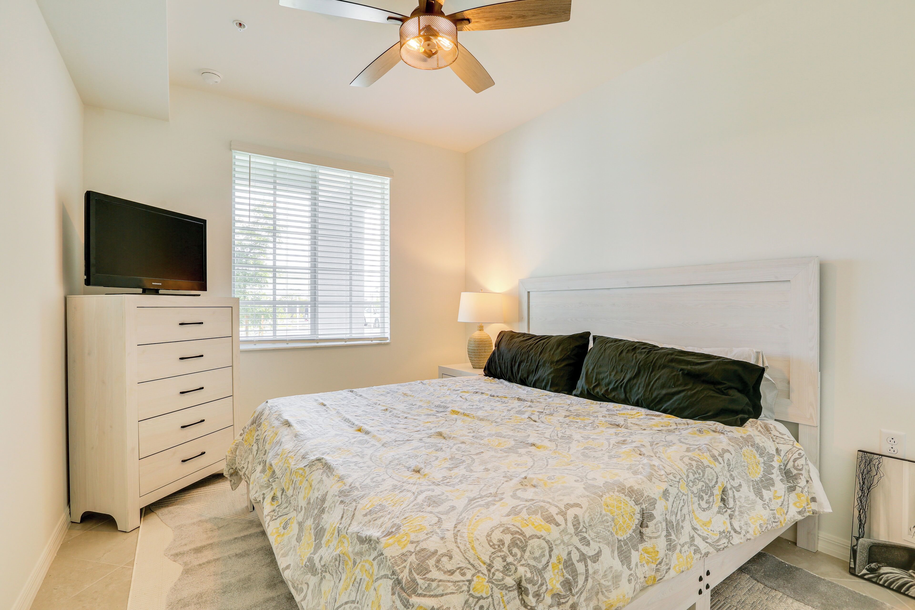 A bedroom with a bed, a dresser, a television on top, a window with blinds, and a ceiling fan.