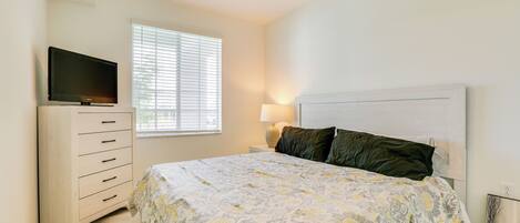 A bedroom with a bed, a dresser, a television on top, a window with blinds, and a ceiling fan.