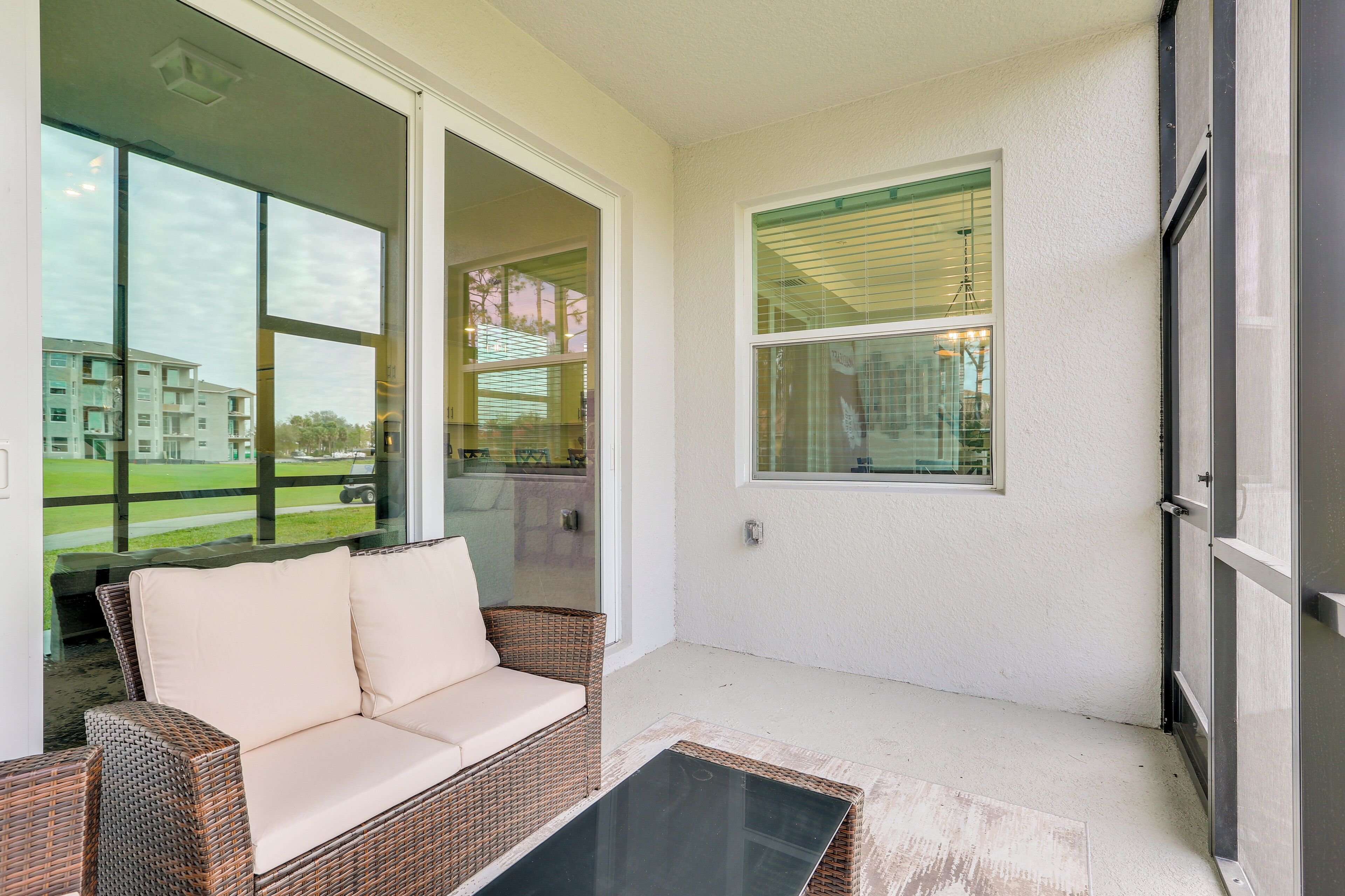 A balcony with a wicker sofa and a glass table, overlooking a building and a grassy area.