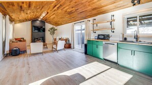 A spacious living area with a wooden ceiling, a fireplace, a dining table, and a kitchen with green cabinets and a window. - Modern Oregon Cottage w/ Hot Tub: Walk to Beach! (Cloverdale)