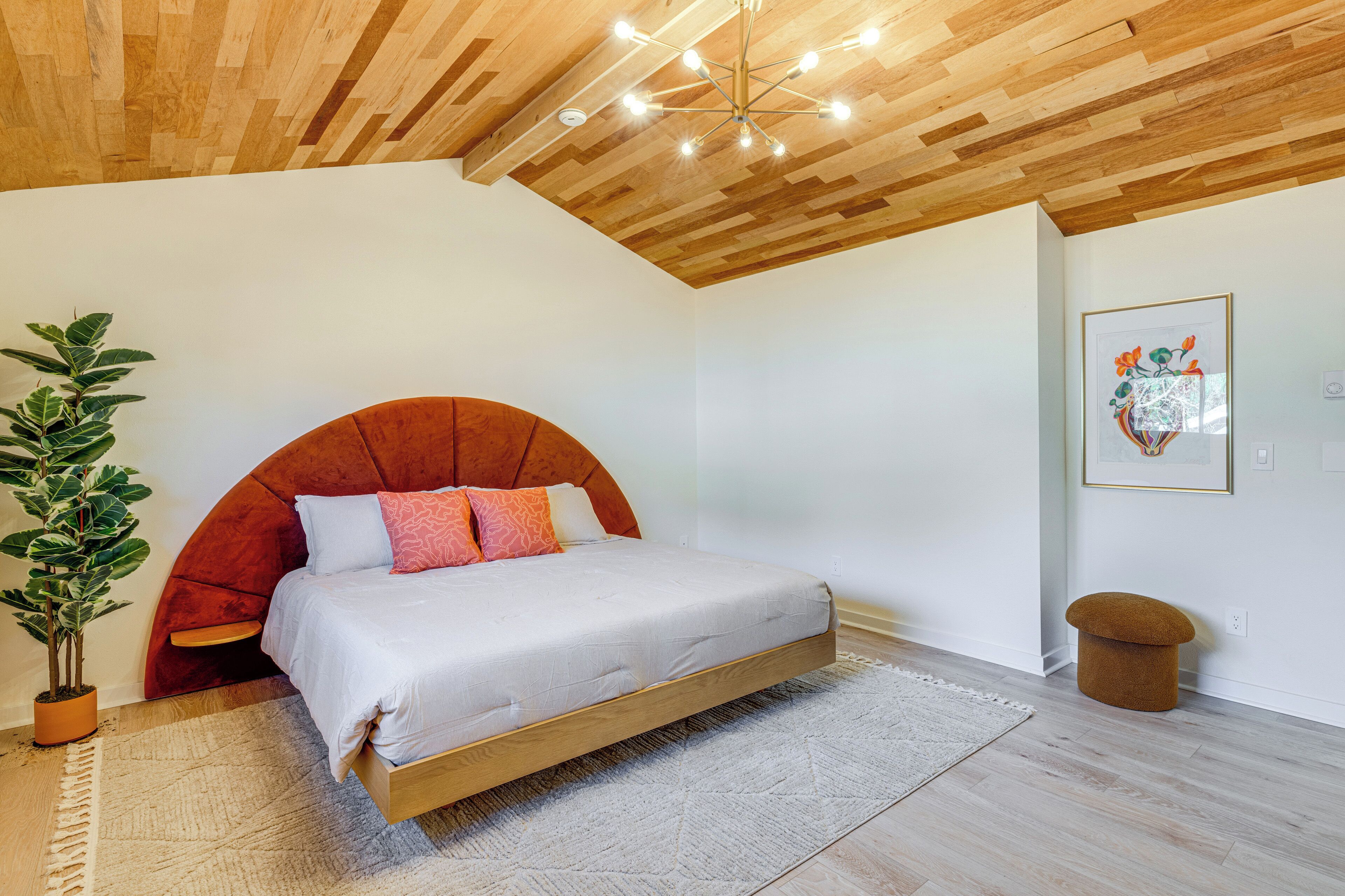 A bedroom with a wooden ceiling, a bed with a curved headboard, a potted plant, and a framed picture on the wall.