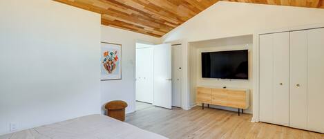 A bedroom with a large bed, wooden ceiling, white walls, and a TV mounted on the wall.