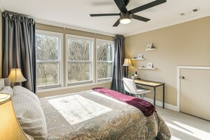 A bedroom with a bed, desk, chair, and large windows.