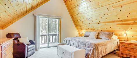 A bedroom with a sloped ceiling, a bed, a dresser, a bench, and a window with a view of trees.