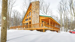 Une cabane en rondins avec une cheminée en pierre, une terrasse en bois et un environnement enneigé.