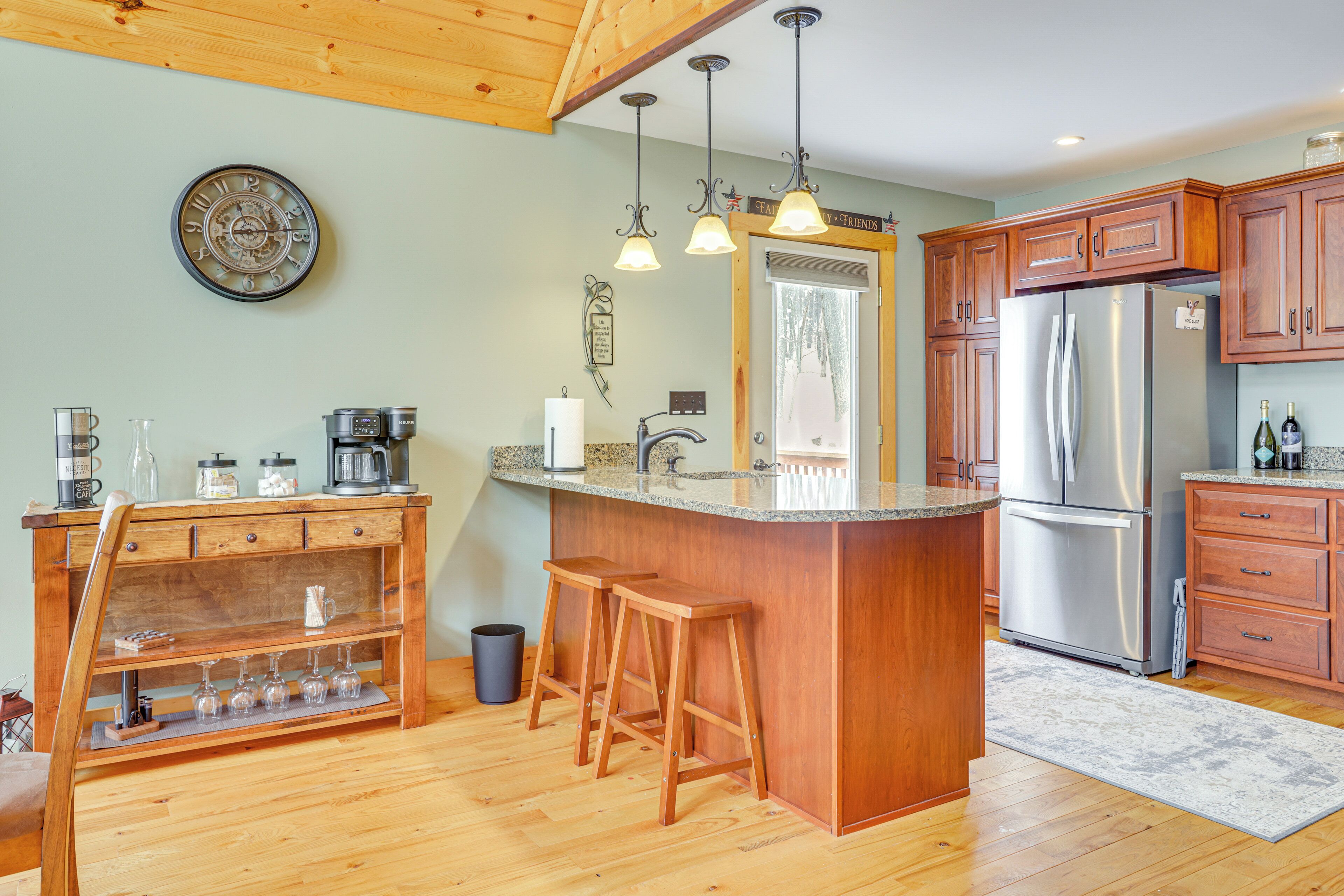Una cocina con armarios de madera, un refrigerador de acero inoxidable, una barra de bar con taburetes y un reloj en la pared.