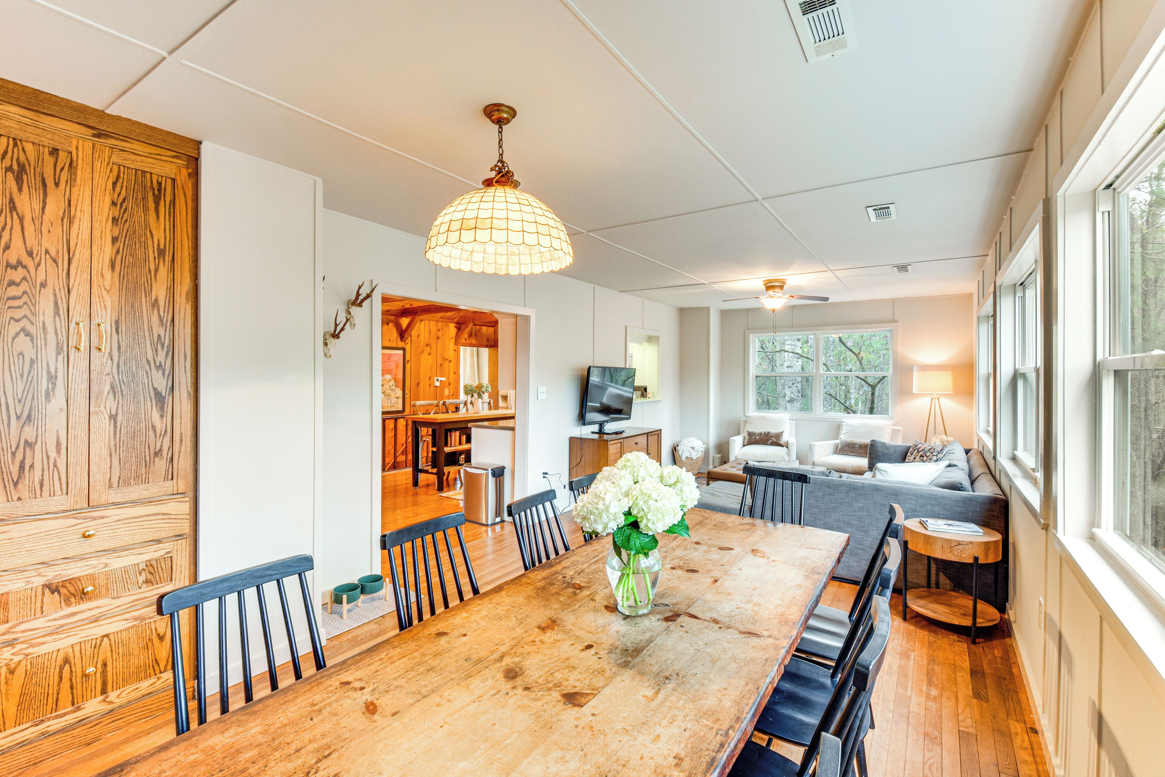 Un grand espace dédié à la salle à manger, avec une table en bois, des chaises et un vase rempli de fleurs. On aperçoit un canapé et une lampe au fond.