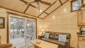 A cozy cabin interior with a wooden ceiling, a sofa with decorative pillows, a wooden coffee table, and a view through a sliding glass door to an outdoor deck. - Minong Flowage Cabin w/ Fire Pit & Dock! (Gordon)