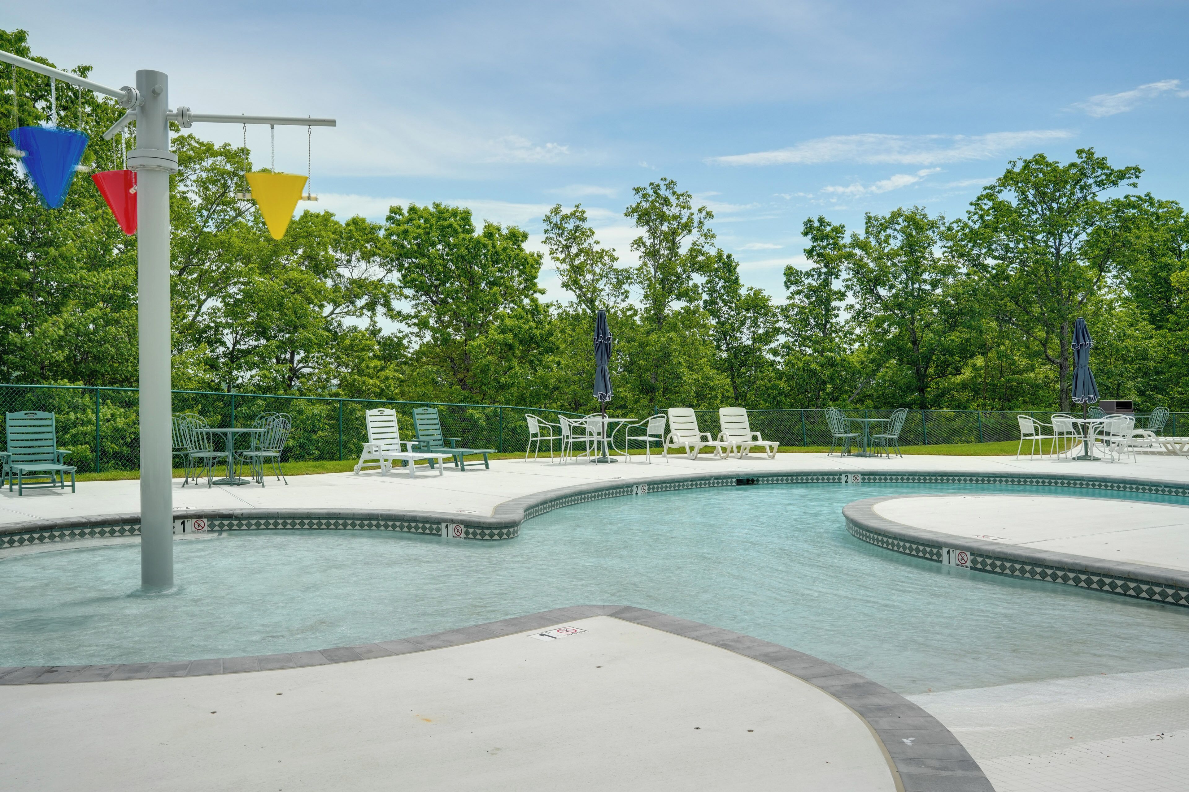 A swimming pool with a tiled edge, surrounded by green chairs and tables, and a metal fence.