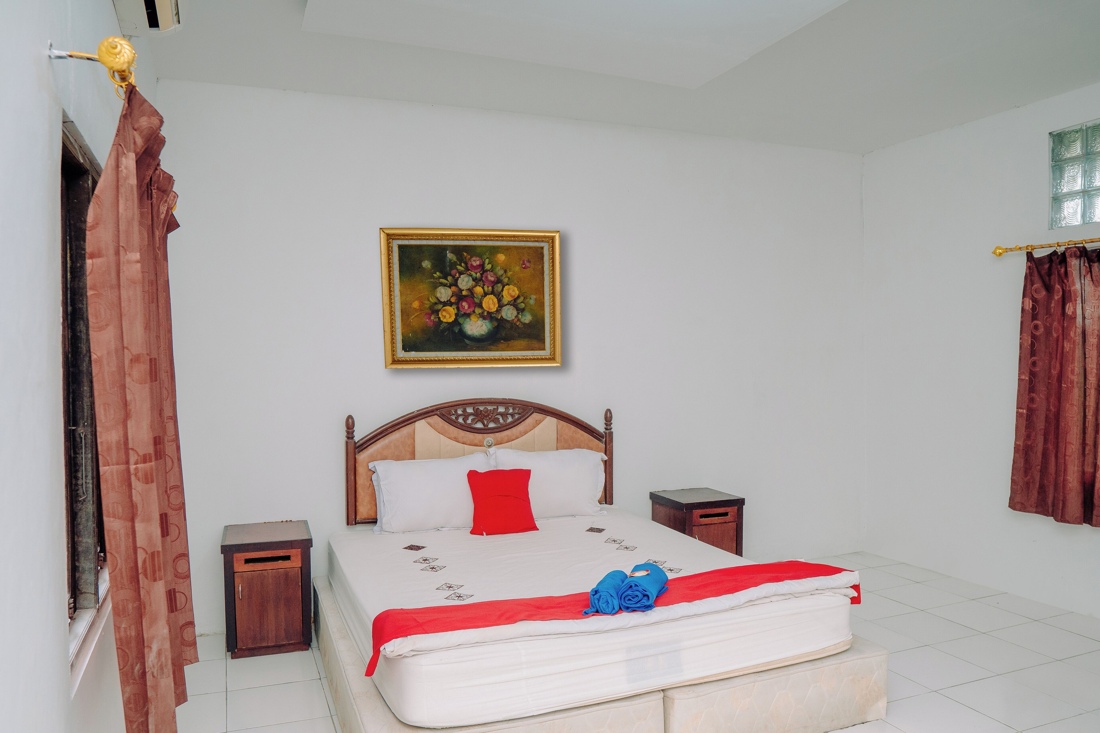 A neatly made bed with a red pillow, a wooden headboard, and a painting of flowers on the wall.