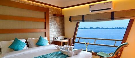 A hotel room with a large bed, a wooden headboard, a window with a view of the sea, and a ceiling fan.