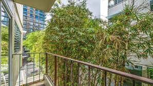 Een balkon met een metalen hekwerk, omringd door dichte bamboe begroeiing, met uitzicht op een stadsbeeld met moderne gebouwen.