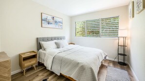 Een slaapkamer met een bed, nachtkastje, vloerlamp, raam met jaloezieën en een schilderij aan de muur.