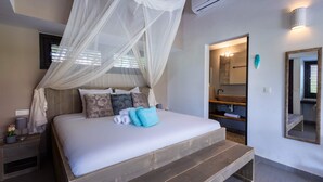 A bedroom with a canopy bed, a wooden bench, a nightstand, a mirror, and a bathroom visible through an open door. - Blue Bay s Tropical Escape (Sint Michiel)