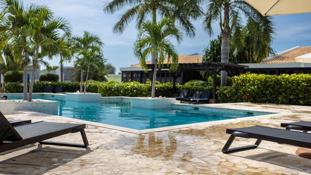 A swimming pool with lounge chairs and umbrellas, surrounded by palm trees and a building with a tiled roof. - Blue Bay s Tropical Escape (Sint Michiel)