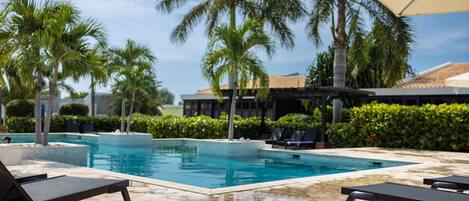 Uma piscina com espreguiçadeiras e guarda-sóis, cercada por palmeiras e um edifício com telhado de azulejos.