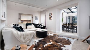 A modern living room with a white sofa, a round coffee table, and a cowhide rug. There is a kitchen area in the background with white cabinets and a dining area with wooden chairs. - Rosemount X3 Bed Townehouse With Hot Tub (Windermere)