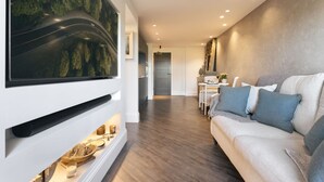 En moderne stue med et stort fladskærms-TV på væggen, en sofa med puder, et spiseområde med bord og stole, og et køkken i baggrunden.