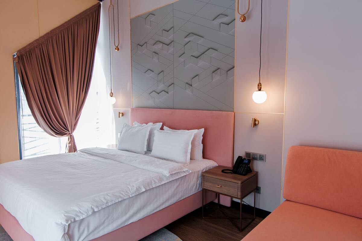 Quarto moderno com cama rosa, roupa de cama branca, mesa de cabeceira com um telefone e painel de parede geométrico.