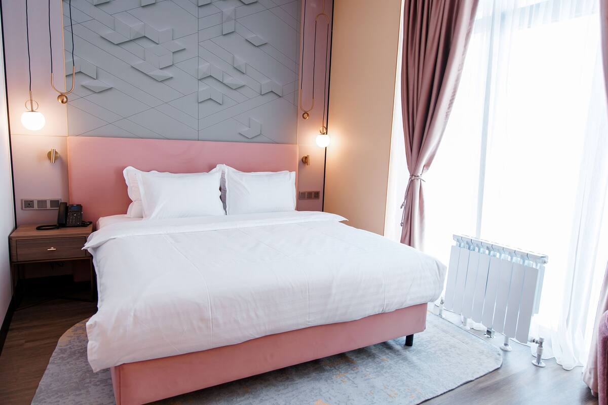 Cama bem arrumada com lençóis brancos, cabeceira rosa e parede cinza com padrão. Há duas luminárias de cabeceira e uma mesa de cabeceira com um telefone e um controle remoto.