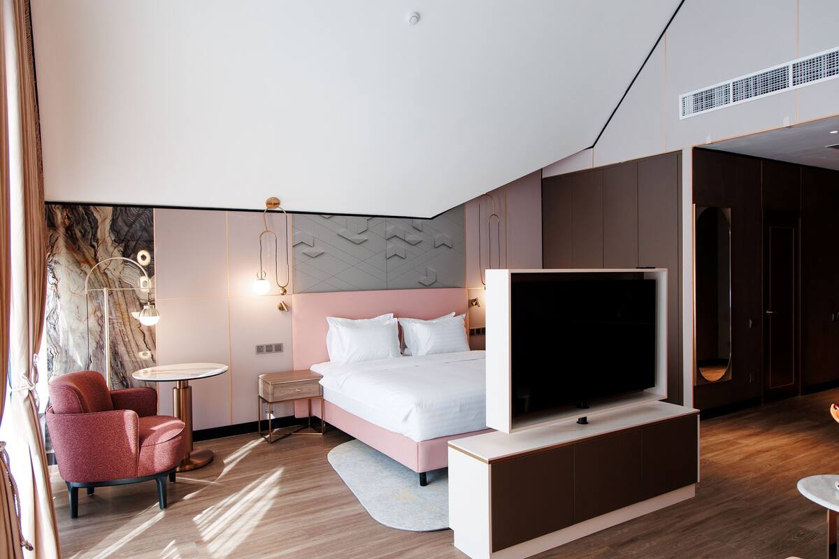 Quarto de hotel moderno com uma cama grande, televisão de tela plana, poltrona vermelha, mesa redonda pequena e parede com padrão marmorizado.