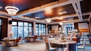 Un amplio salón de banquetes con varias mesas dispuestas para comer, grandes ventanales que ofrecen vistas y modernas lámparas.