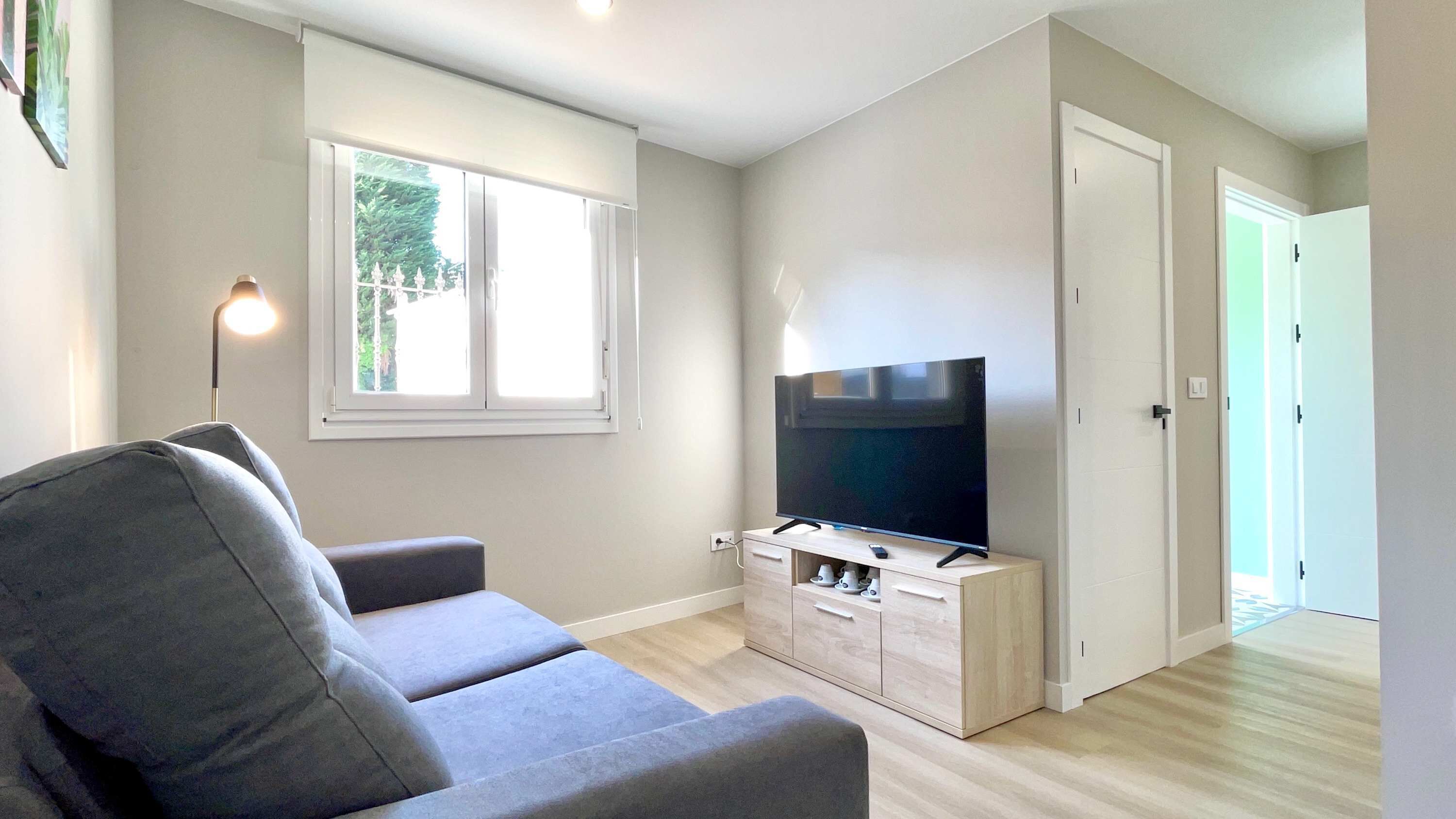Una sala moderna con un sofá gris, un televisor de pantalla plana sobre un mueble de madera, una ventana con persianas y una puerta que da a otra habitación.