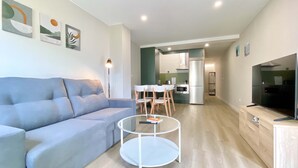 Uma sala de estar moderna com um sofá cinzento, uma mesa de centro de vidro e uma televisão. Há uma área de cozinha com uma mesa de jantar e cadeiras.
