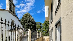 Um corredor com chão de azulejos, um edifício branco com uma torre e uma árvore ao fundo.