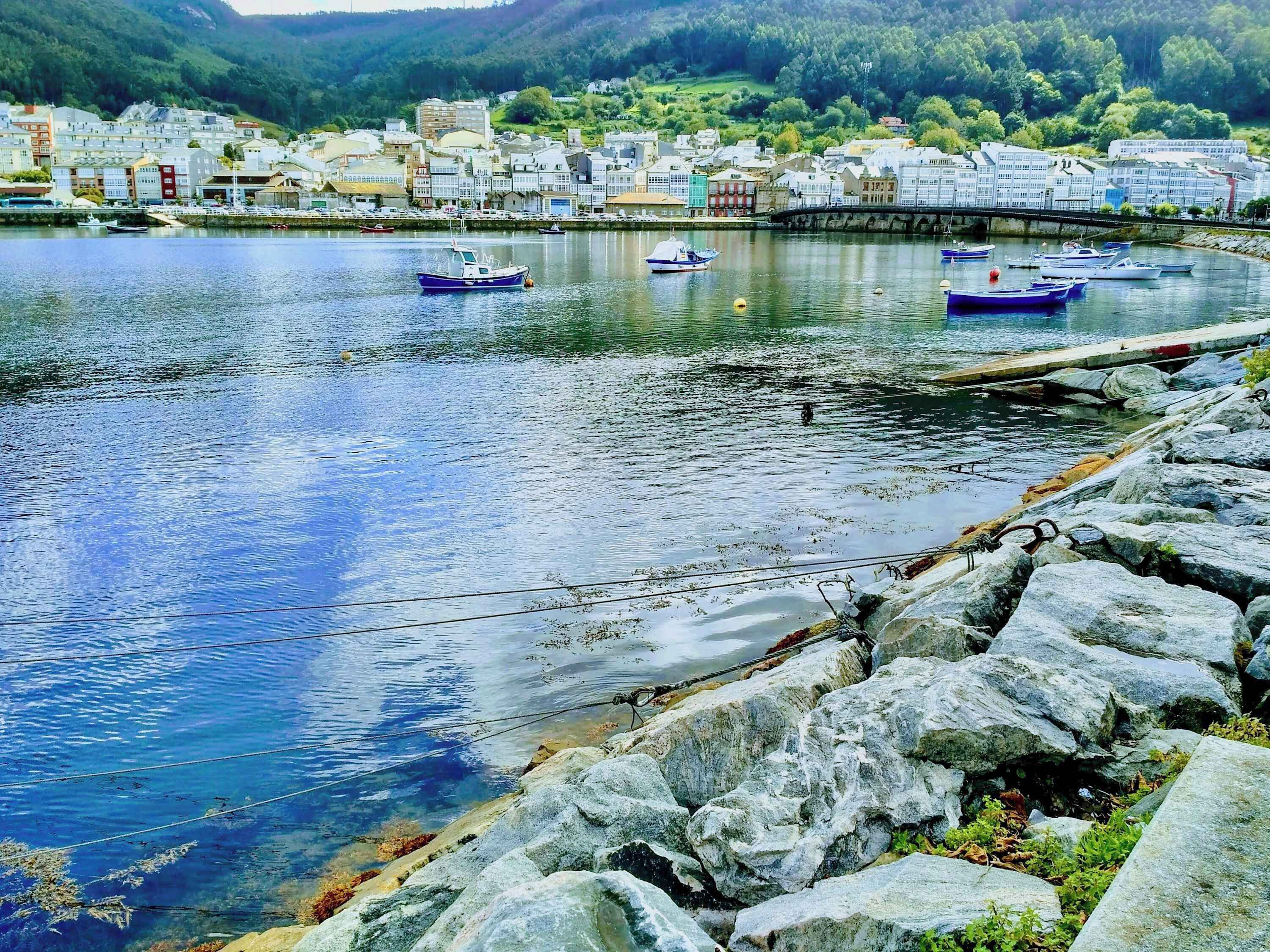 A waterfront town with boats docked, rocky shore, and buildings in the background.