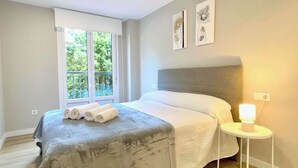 A bedroom with a bed, a bedside table with a lamp, a window with a view of greenery, and artwork on the wall. - The Cliffs - AT Seiramar Portonovo (Viveiro)