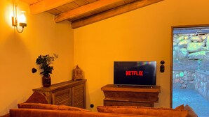 Un salon chaleureux avec un plafond en bois, un téléviseur affichant le logo « NETFLIX », un canapé marron, une table en bois sur laquelle se trouve un pain, et un mur de pierre donnant sur un chemin de pierres.