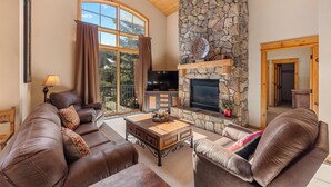 A spacious living room with leather sofas, a stone fireplace, a flat-screen TV, and large windows.