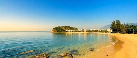 A beach with clear water, rocks, and buildings in the background.