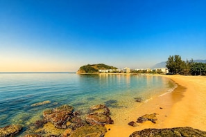 A beach with clear water, rocks, and buildings in the background.