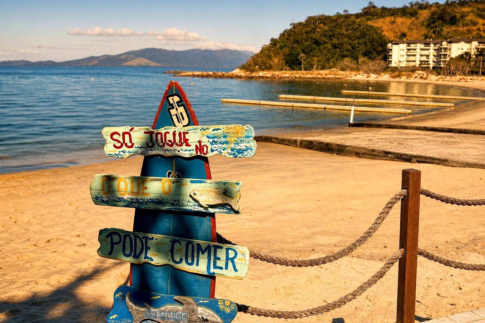 Placa de sinalização à beira-mar com várias indicações de direção, uma barreira de corda e o cenário de uma praia, oceano e montanhas ao longe.