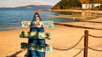 Placa de sinalização à beira-mar com várias indicações de direção, uma barreira de corda e o cenário de uma praia, oceano e montanhas ao longe.